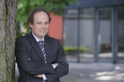 This picture shows Jürgen Renn, one of the directors at the MPI for Geoanthropology in Jena. He is leaning against a tree in front of the institute building, wearing a blue jacket and a blue and grey striped tie. His arms are crossed and he is looking into the camera.